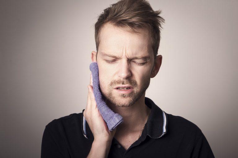 Man wincing while holding a purple towel against his jaw and cheek, showing signs of jaw discomfort
